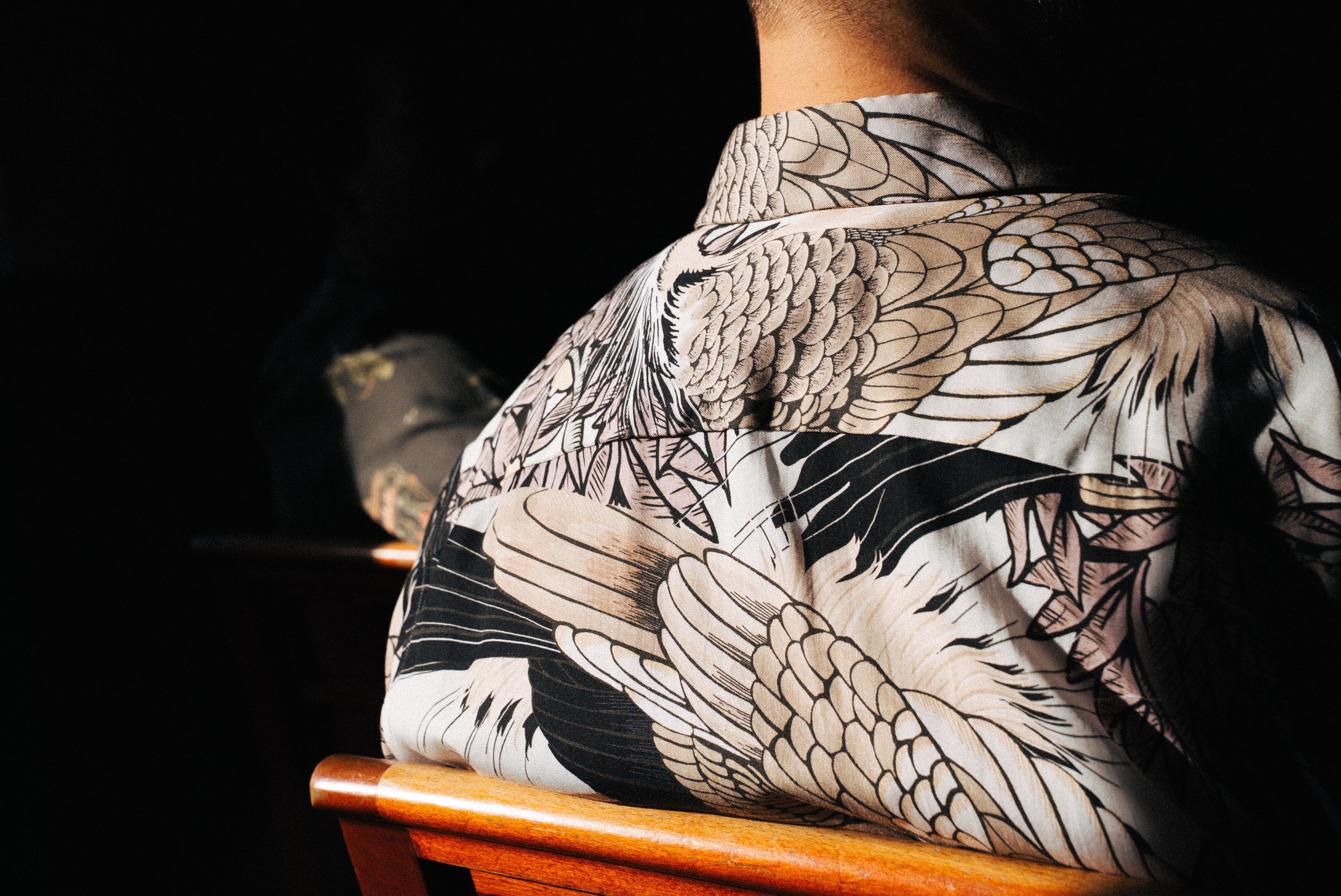 Person wearing a Ronni shirt with a detailed bird pattern sitting on a wooden chair.